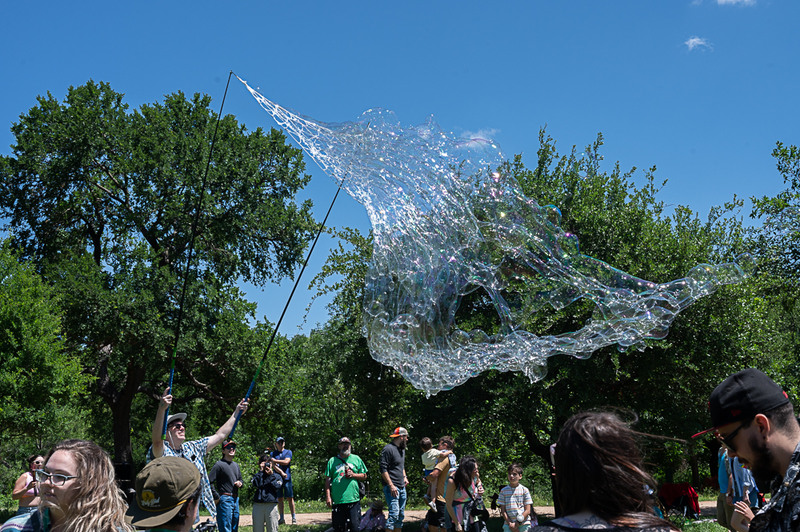 A man creates a giant, iridescent soap bubble using poles and string, entertaining a crowd in a sunny park with trees in the background.