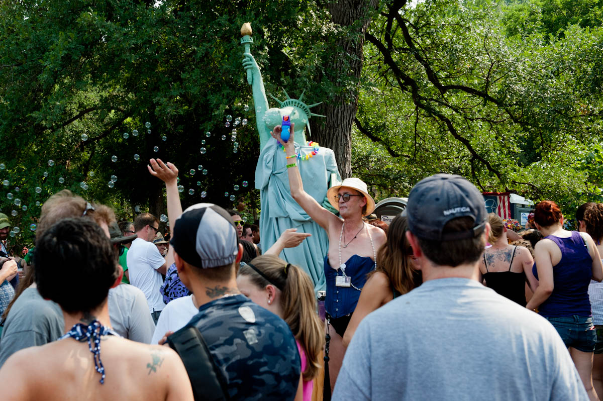 A crowd gathers outdoors under trees, with bubbles floating in the air. A person dressed in a Statue of Liberty costume holds up a small, colorful object. Another individual in a hat and summer attire gestures enthusiastically, adding to the festive atmosphere.